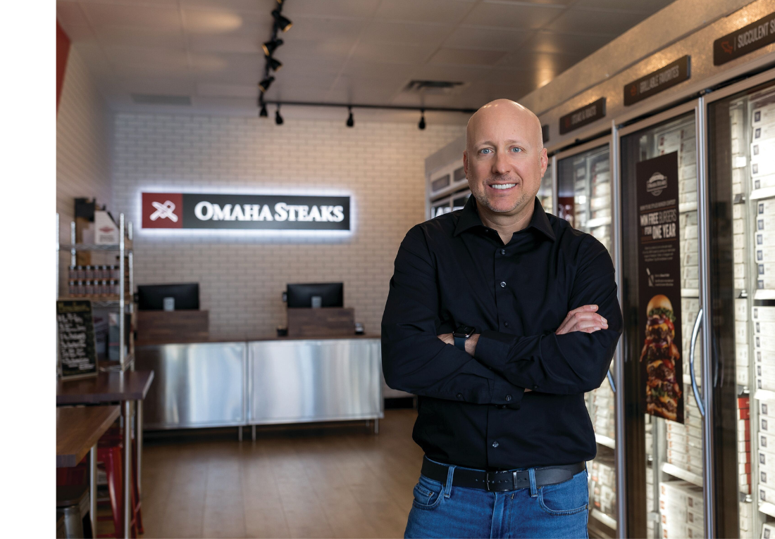 Nate Rempe, President and CEO of Omaha Steaks, standing with arms crossed and smiling inside an Omaha Steaks retail store, with the company logo and freezer section visible in the background.