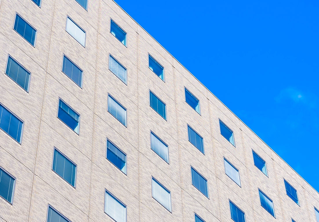 Exterior view of a modern building with a beige facade under a clear blue sky. The building has a geometric design with rows of windows reflecting the bright sunlight.