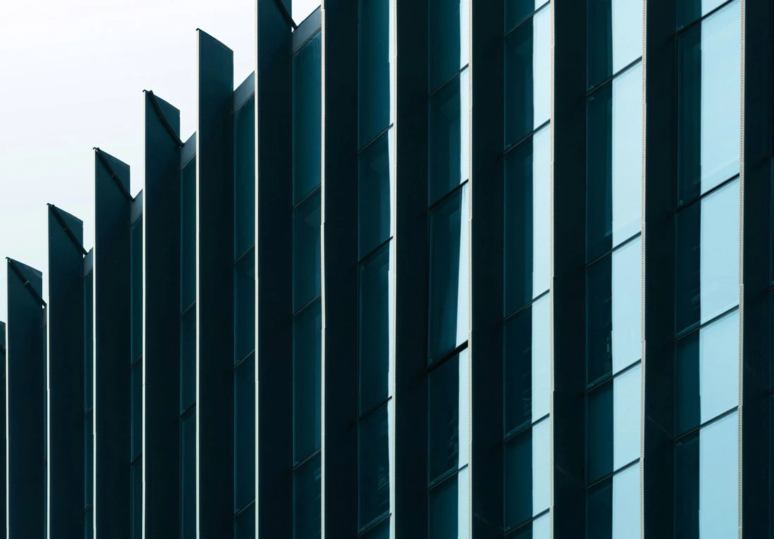 Close-up view of a modern building facade featuring vertical, evenly spaced, dark-colored fins and large glass windows reflecting the sky.