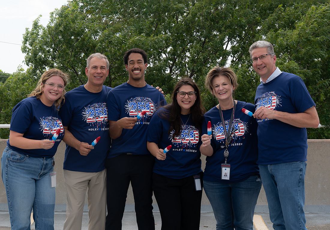 Six Koley Jessen employees wearing commemorative 250th anniversary T-shirts and holding red, white, and blue popsicles outdoors.