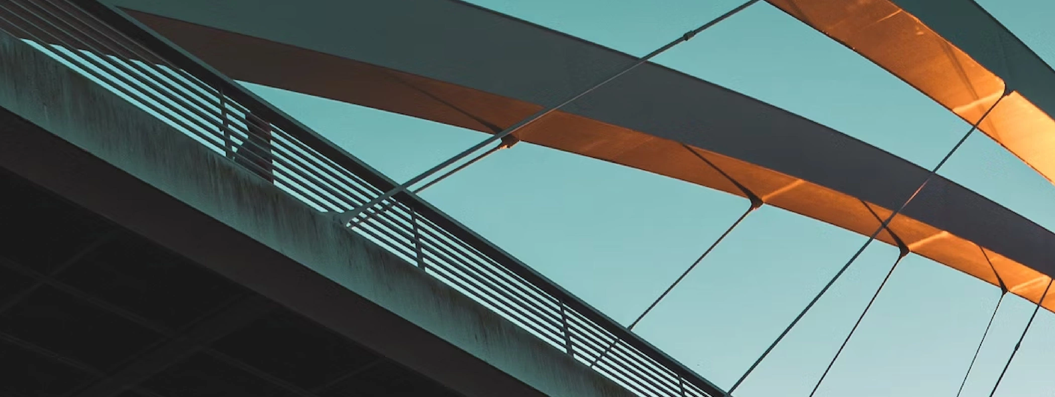 A close-up view of a modern bridge against a clear sky. The bridge features a sleek, curved design with an underside illuminated by warm sunlight, creating a contrast of light and shadow. The railing and cables are visible, adding to the architectural det